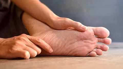 Fuß eines asiatischen Mannes: Zwei Hände massieren die Fußsohle eines nackten Fußes, der auf einem Holzboden aufliegt.