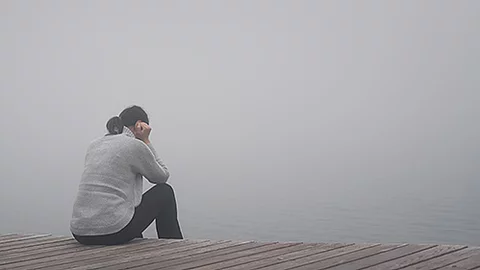 Eine Person sitzt auf einem Holzsteg am Wasser, umgeben von dichtem Nebel. Die Haltung wirkt nachdenklich und zurückgezogen